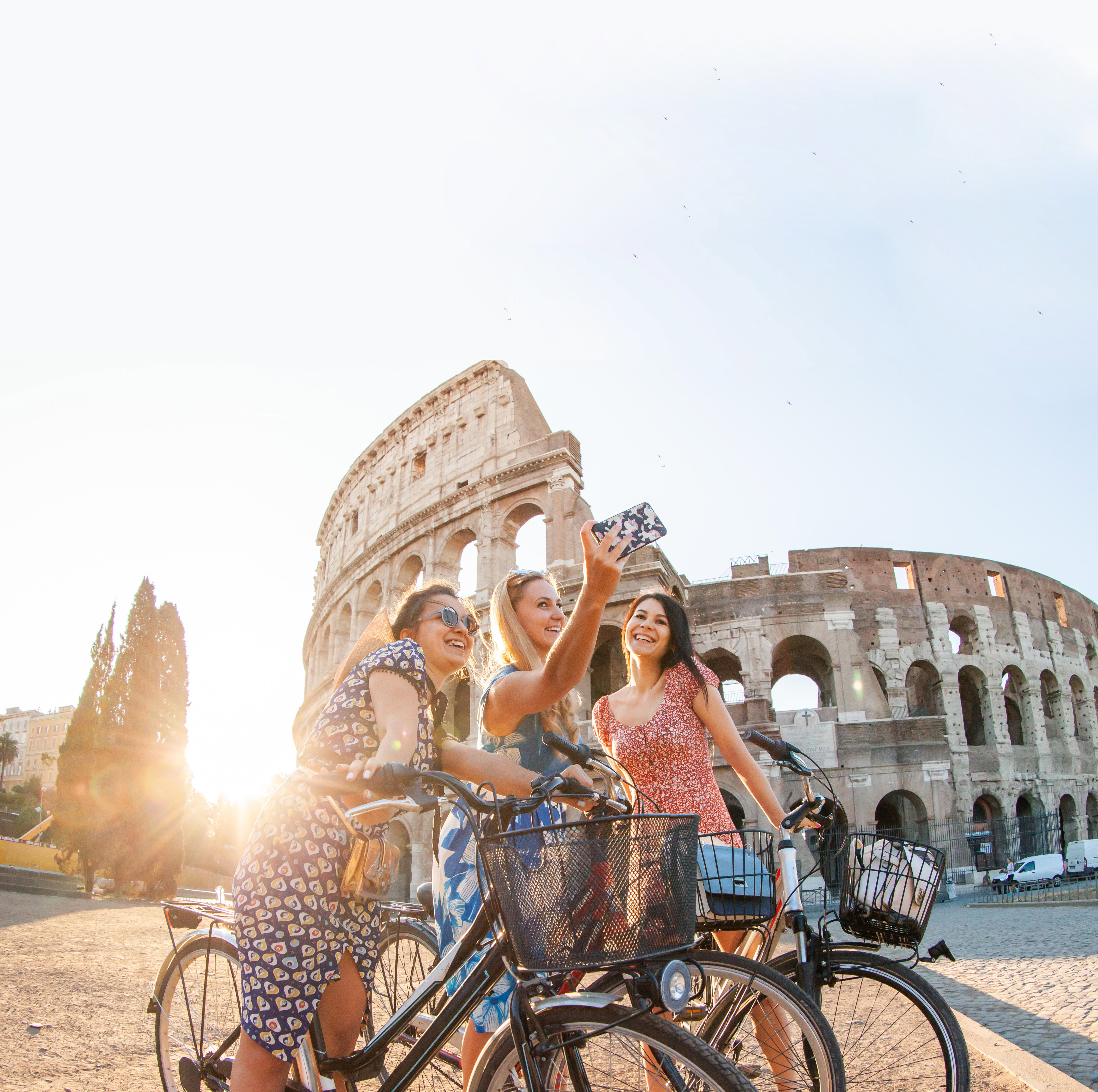 Three women wearing sun dresses taking a selfie from their bikes in front of the Colosseum at sunrise, Rome, Italy.