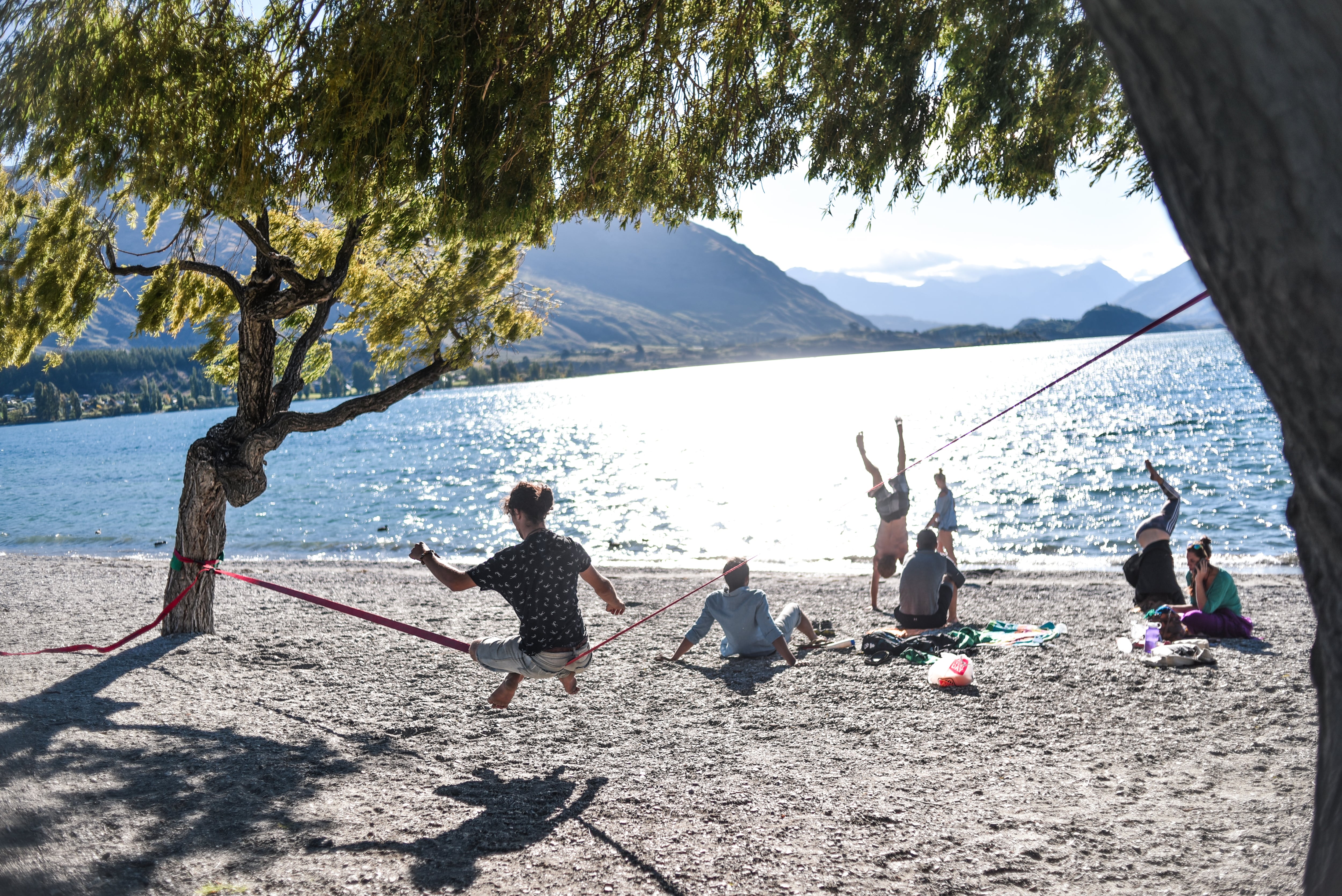 A group of travellers hang out on Wanaka Beach looking towards the water glittering in the sunlight.