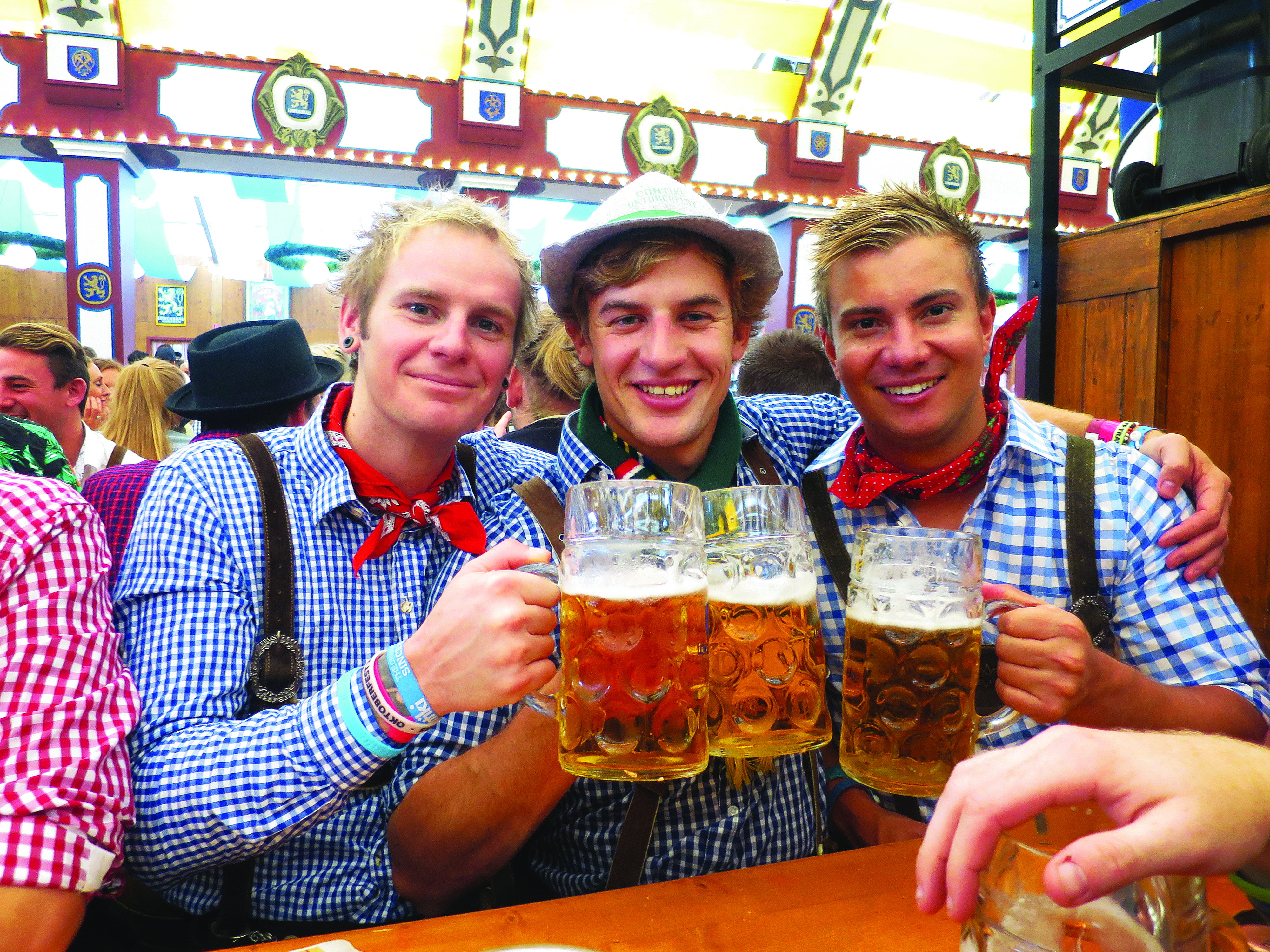 Three men smile for the camera wearing traditional blue check shirts and laderhosen while cheering with their beer steins at Oktoberfest, Munich, Germany
