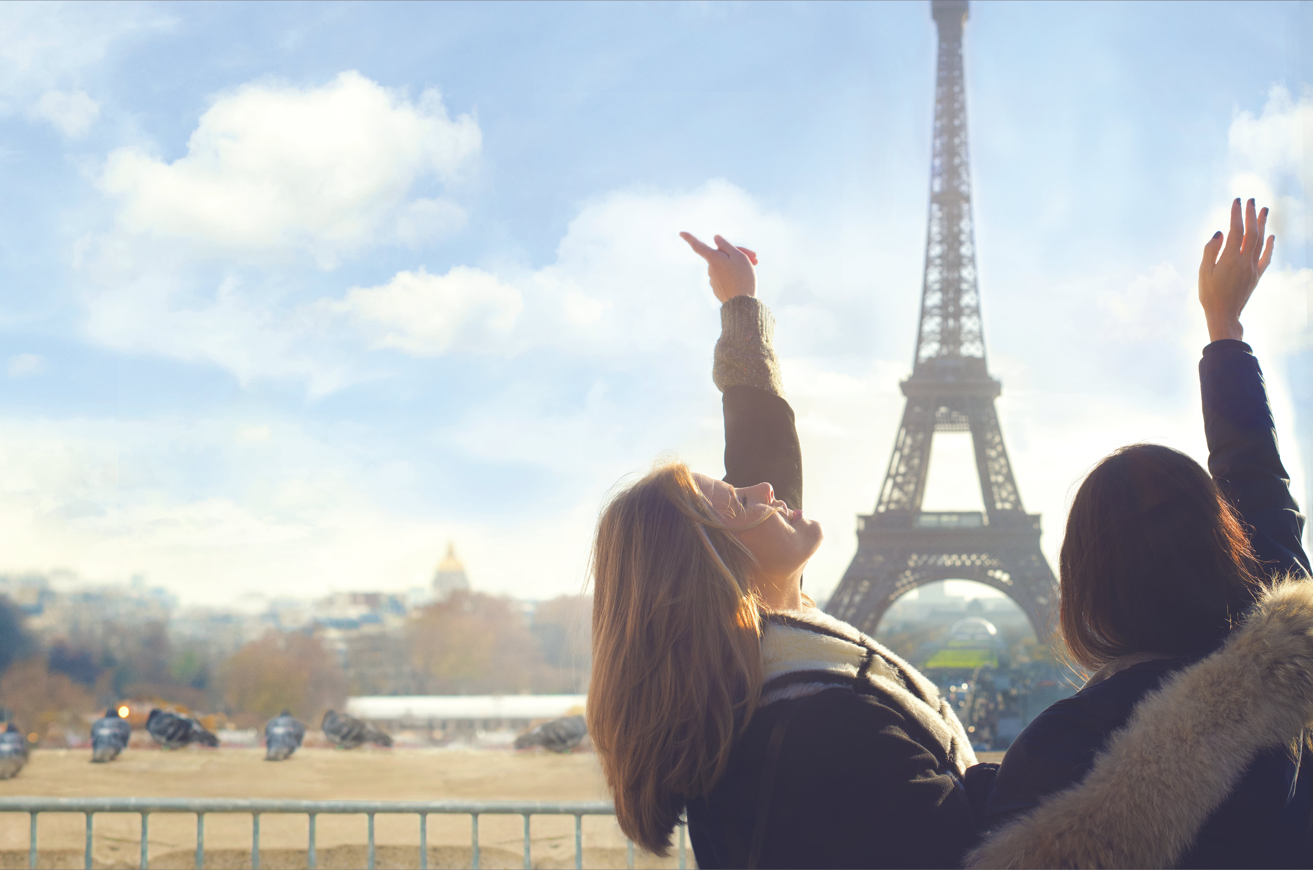Two female friends wearing winter coasts and scarves sing with their hands in the air overlooking the Eiffel Tower at sunset, Paris, France