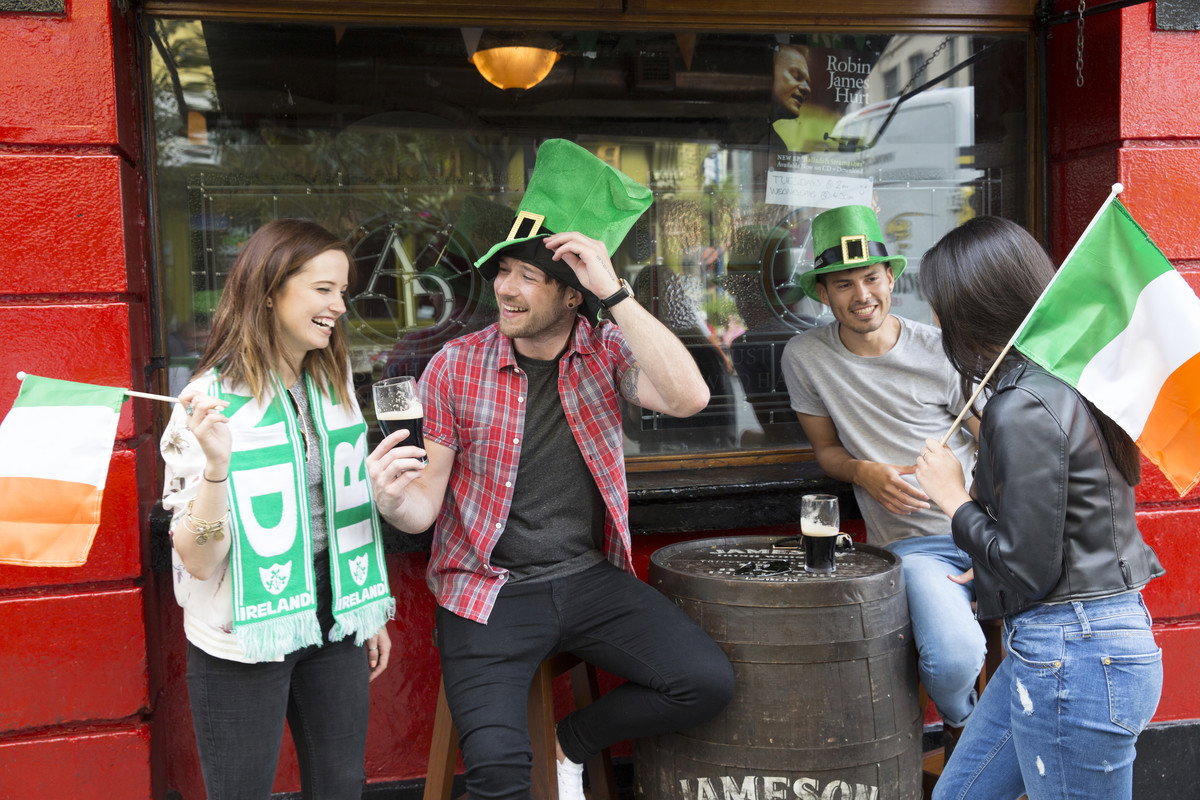 A group of young friends wearing green top hats and waving Irish flags enjoy a pint of Guinness outside of a pub in Dublin on St Patrick's Day, Ireland