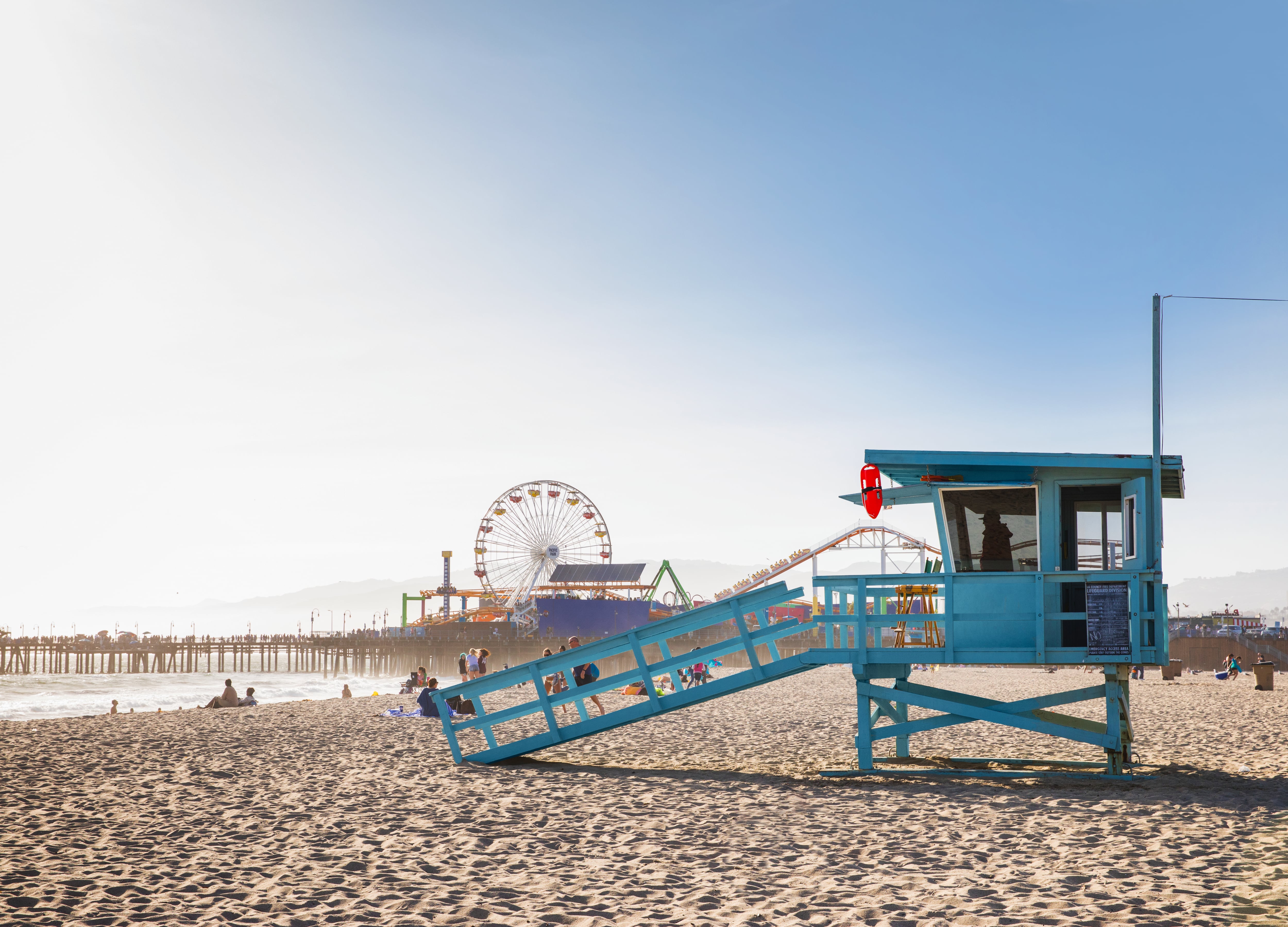 a colourful turquoise lifeguard tower sits on the wide sand of Santa Monica Beach, in the background is the Santa Monica Pier with its ferris wheel and wooden rollercoaster