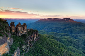 A wide angle shot of the Blue Mountains, with the Three Sisters rock formations stood in front of a pink sky at sunset, Australia
