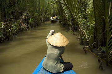 People are rowed down the murky brown waters of the Mekong River between tall aquatic grasses on bamboo rafts