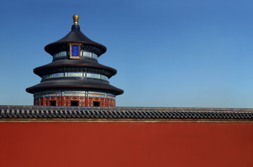 The top of a tiered red temple stands above the roofs of ancient buildings set against a clear blue sky in Beijing, China