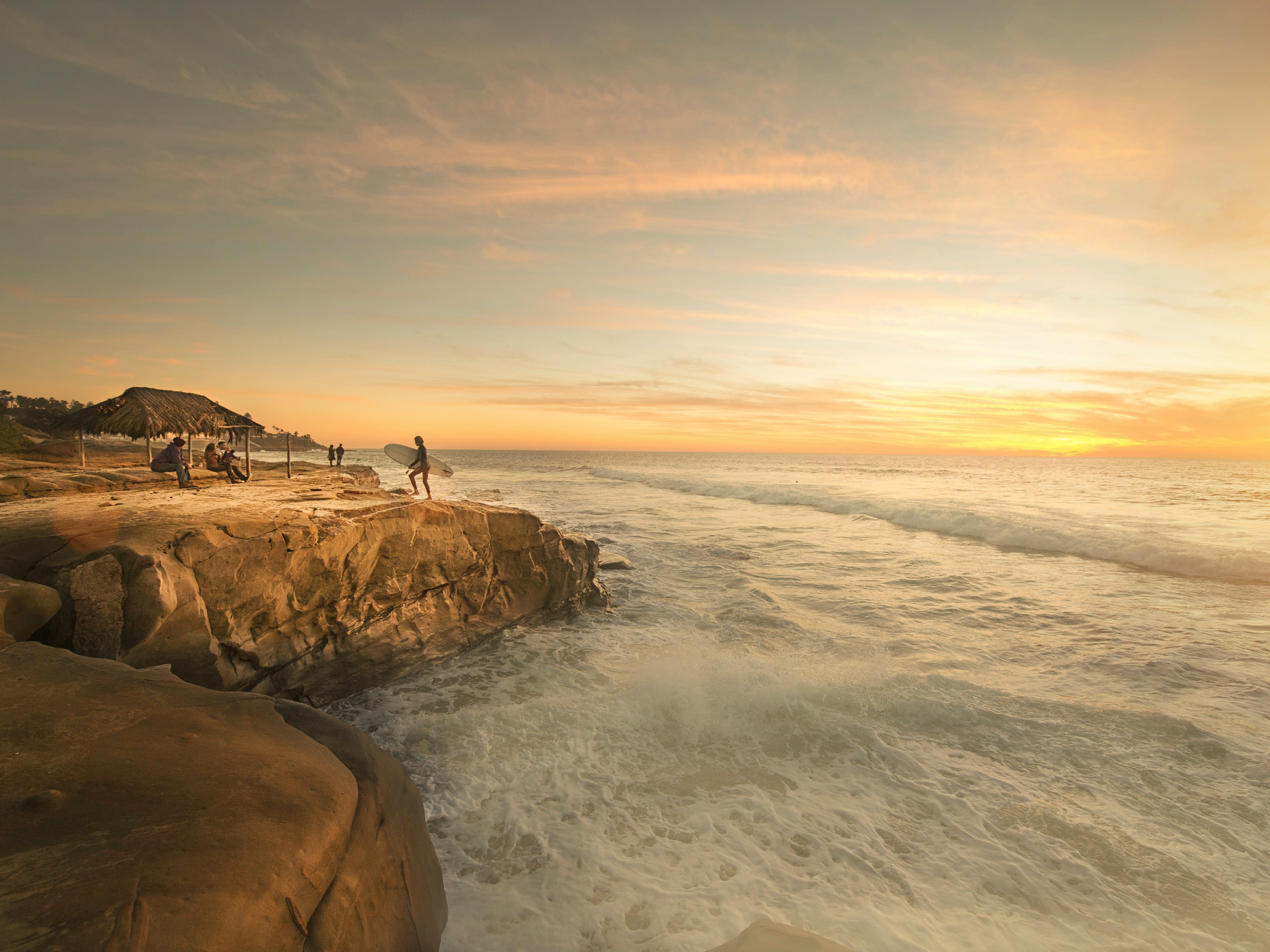 A hazy sunset over the Pacific Ocean in San Diego, California. Waves break against a rocky shore and a surfer stands on the shore holding his surfboard