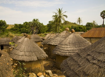 Deep orange, earthen huts with thatched roofs stand close to one another with a treeline and rolling clouds in the background, Tamberma Valley, Togo, Arica