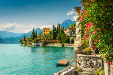 A small town of Venetian style buildings, cypress trees and a church with a tower sit on Lake Como, verdant mountains tower in the distance, Italy