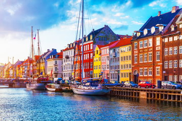 Vibrant coloured buildings of yellows, reds, oranges and blues line the harbour, where small colourful sail boats bob in the water on a bright sunny day. Copenhagen, Denmark