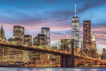 The New York City skyline lit up at dusk behind the Brooklyn Bridge underneath a pink and blue sky at sunset.
