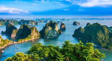 The limestone karsts of Halong Bay Vietnam tower over the blue water with small junk boats and blue sky.