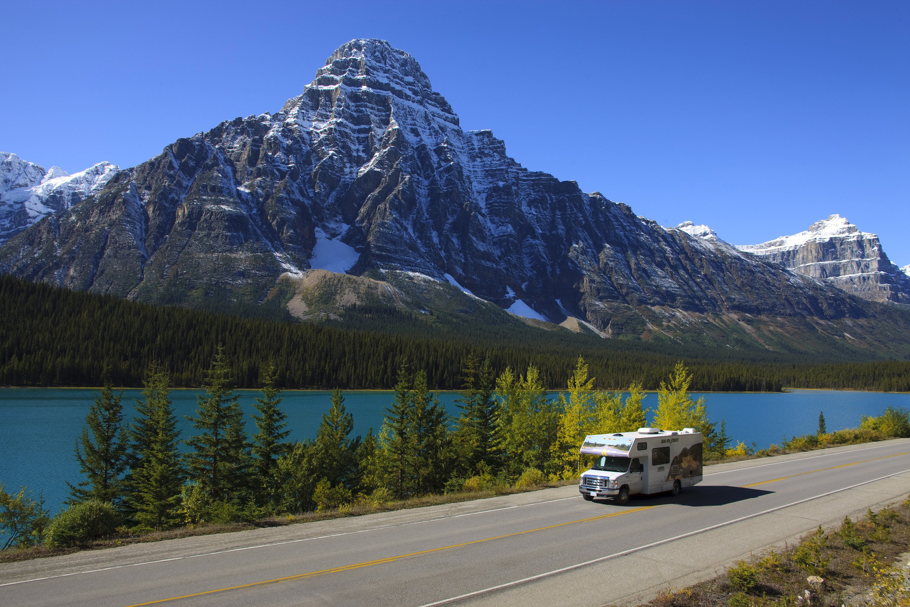 A Cruise Canada campervan drives among lush green trees with a lake and mountain backdrop
