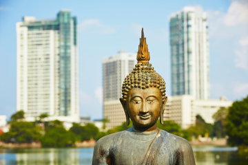 A Buddha statue stands in the foreground in front of a lake with high rise towers in the background in Colombo, Sri Lanka.