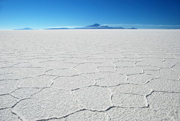 An expanse of dazzling white, hexagonal salt pans sit against a horizon of bright blue sky and volcanos at the Salar de Uyuni, Bolivia
