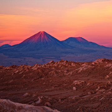 The red rocks at Piedras Rojas on the edge of a light blue lagoon with two volcanoes rising beyond the water with a perfect reflection in the blue waters in the Atacama Desert, Chile