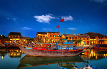 A battered red and blue boats sits on the placid river in Hoi An, Vietnam where  brightly lit yellow French Colonial buildings break the skyline against a dusky sky