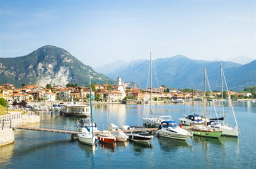 Small boats are moored to a wooden jetty on the beautiful reflective Lake Maggiore, the buildings of the town cling to the bay in the distance with the Alps soaring up behind, Italy