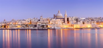 A view of the historic skyline of Valletta and the dome of the Basilica of Our Lady of Mount Carmel lit up and glowing in the purple haze of the sunset in Malta.