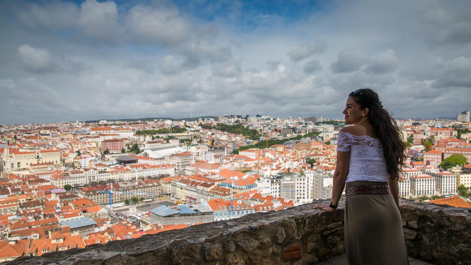 A female traveller with long flowing brown hair leans on a stone wall as she enjoys the view over Lisbon's terracotta roofs.