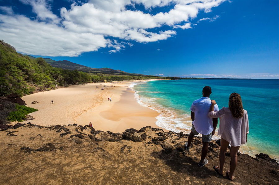 A couple stand on a clifftop staring down at a beautiful white sand beach in Hawaii