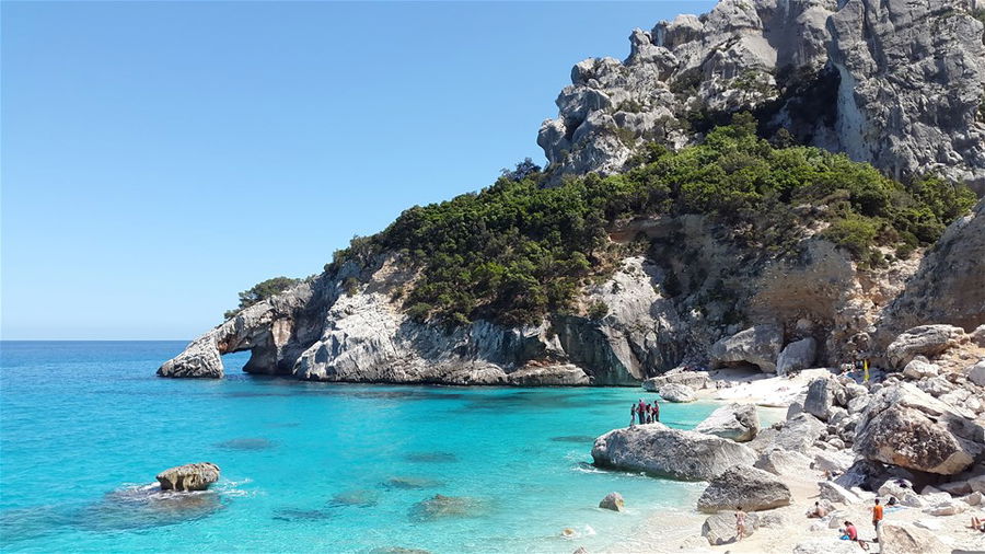 People stand on a rocky beach in Sardinia on a sunny day