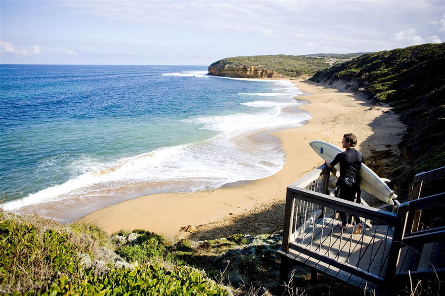 A surfer stands at the top of a wooden staircase which descends down a cliff face to a beautiful beach below in Australia