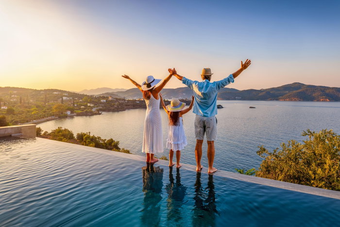 A happy family on summer holidays stands by the swimming pool and enjoys the beautiful sunset over the mediterranean sea in Greece