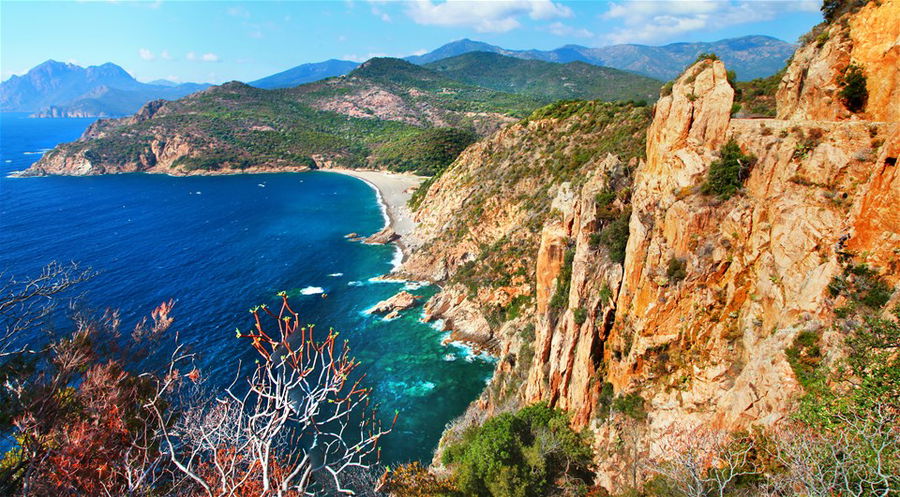 Aerial view of a distant beach with the bright blue ocean, surrounded by steep cliffs, in Corsica France