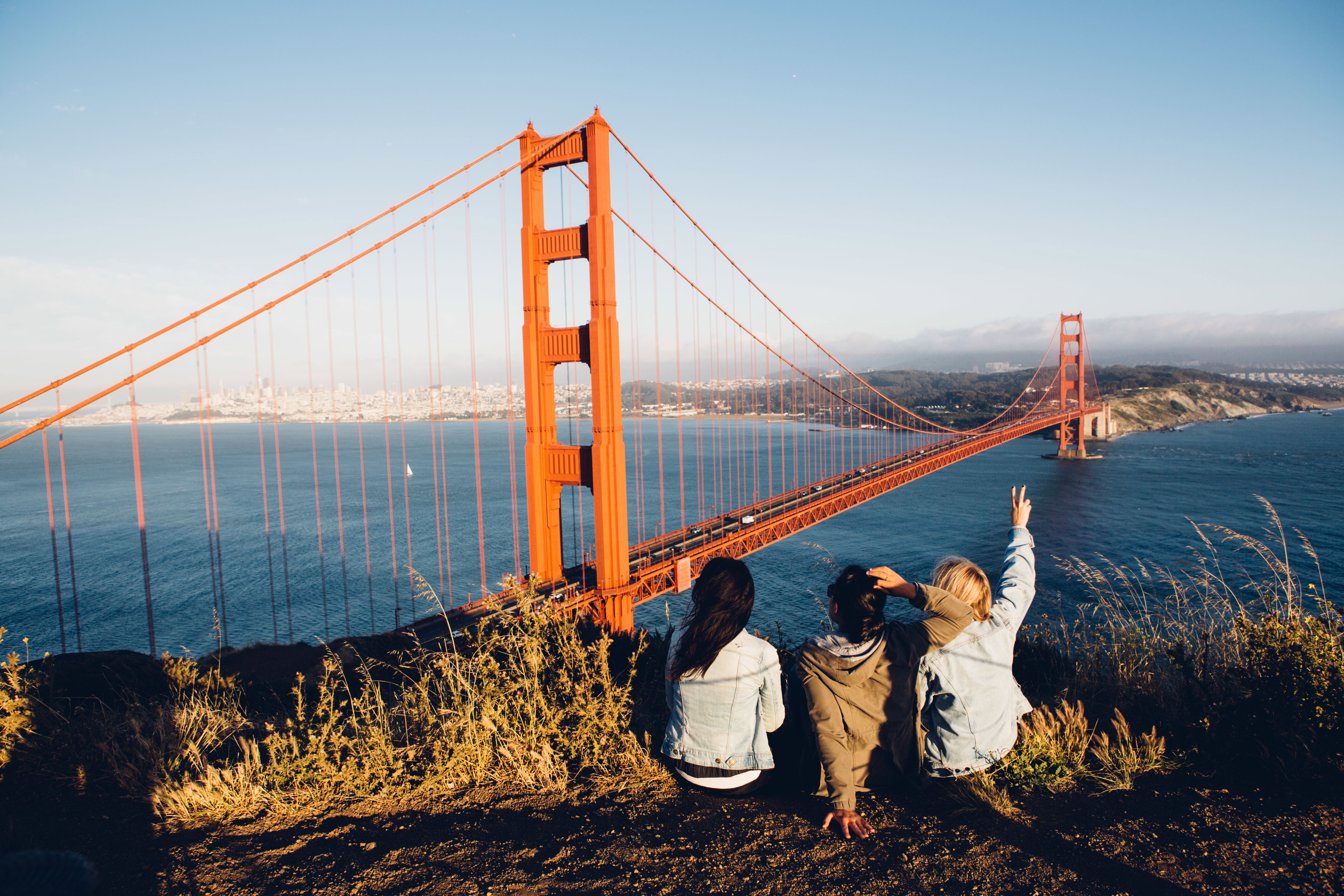 Three young women sit on a bluff, with their backs to the camera, looking out at the view of the Golden Gate Bridge with San Francisco in the background.