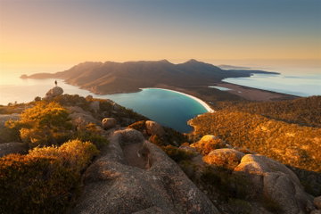 A person stands on a cliff top looking out to the sea and rugged hills in Freycinet National Park, Tasmania