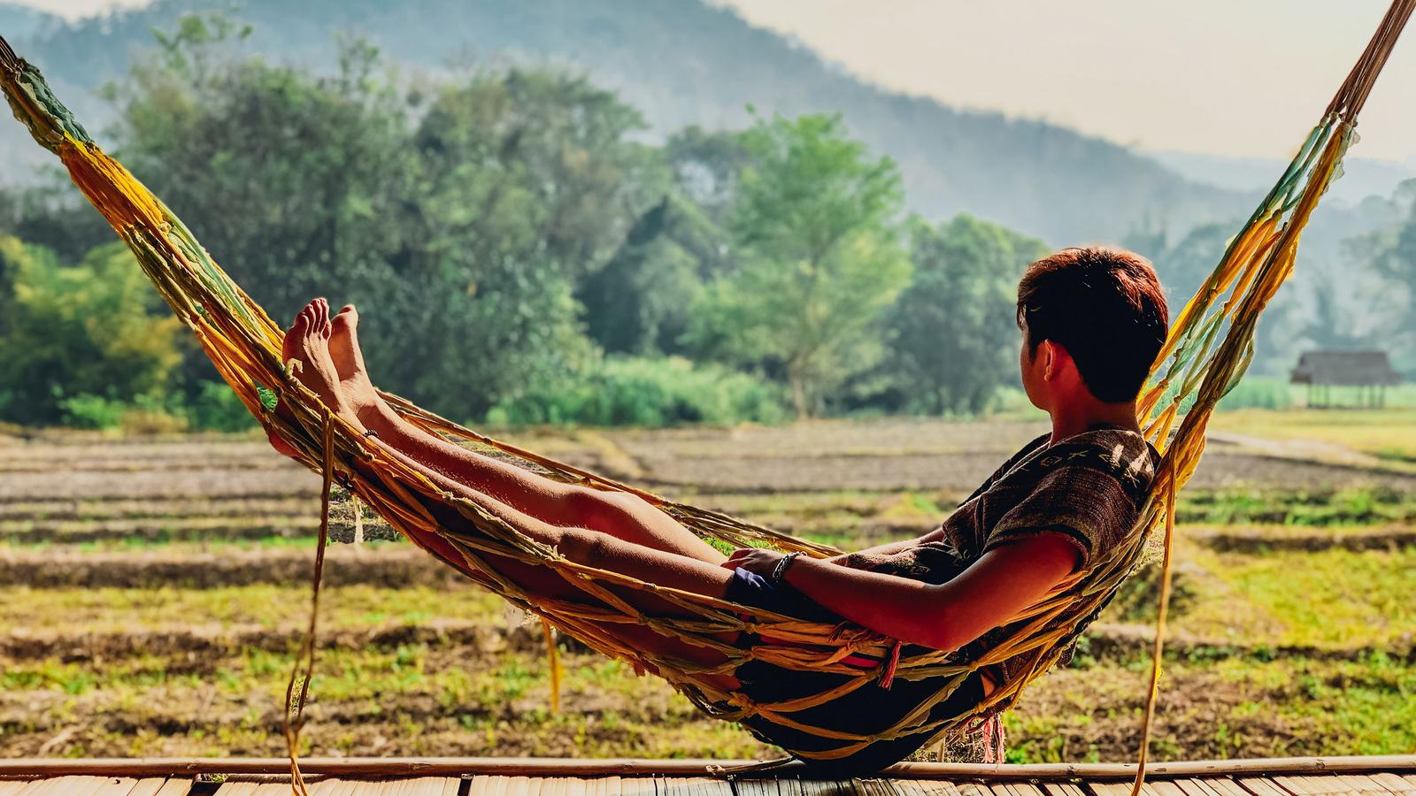 A young man lies in a string hammock in the countryside of northern thailand, looking towards the distant mountains.
