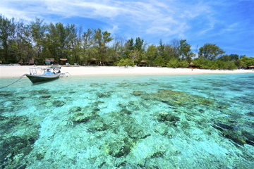 Crystal clear waters surround the white sand and palm fringed island of Gili Trawangan, with a fish boat on the shoreline in the Gili Islands, Indonesia.