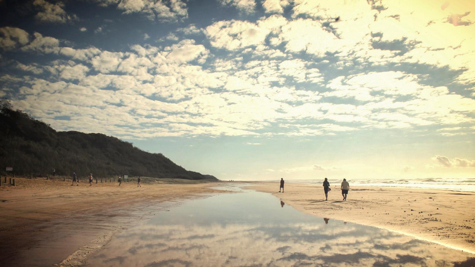 Clouds scud across the sky and are reflected in the wet sands of Fraser Island where people walk along the beach
