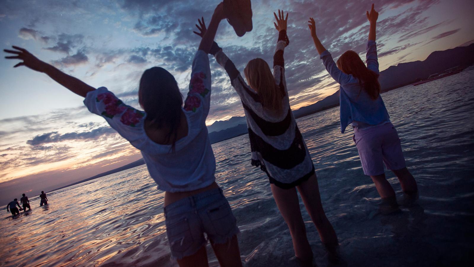 Three women stand in shallow waters with their arms held high at sunset in the Gili Islands, Indonesia